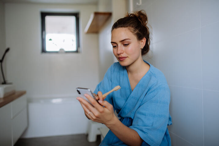 Woman in bathroom brushing her teeth and looking at her phone
