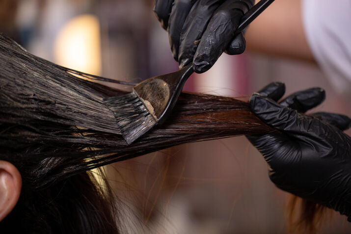 Person with black gloves applying hair dye to a thick strand of hair using a brush.