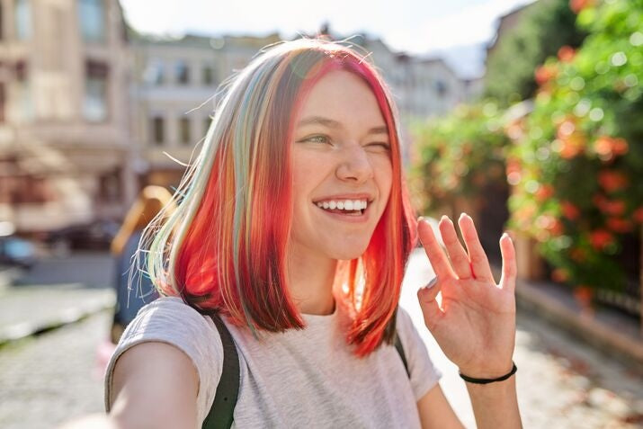 Woman with colorful hair waving on a street, winking with her right eye